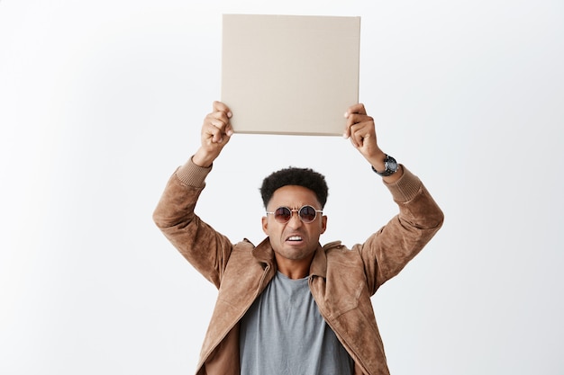 Uno de cada diez. Eww. Retrato de joven atractivo hombre de piel oscura con peinado afro en ropa casual y gafas de sol con cartón vacío arriba con expresión de disgusto.