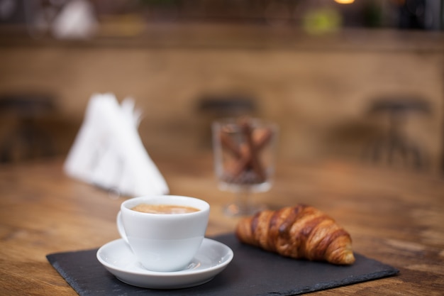 Café capuchino en una taza blanca sobre una mesa de madera junto a un delicioso croissant. Snak sabroso. Cafetería vintage.