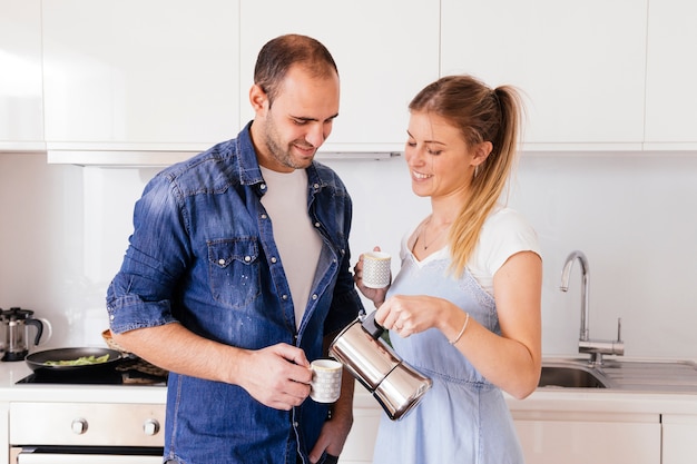 Café de colada sonriente de la mujer joven en control de la taza de su novio en la cocina