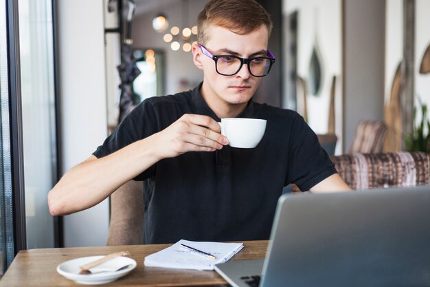 Café de consumición del hombre joven en la tabla con el ordenador portátil