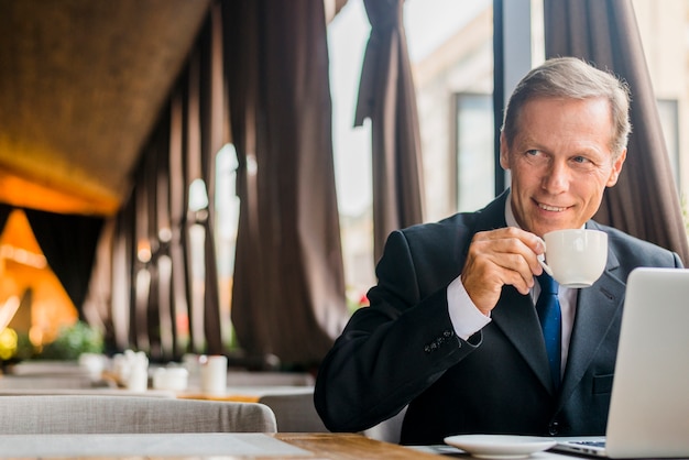 Foto gratuita café de consumición del hombre de negocios feliz con el ordenador portátil en el escritorio