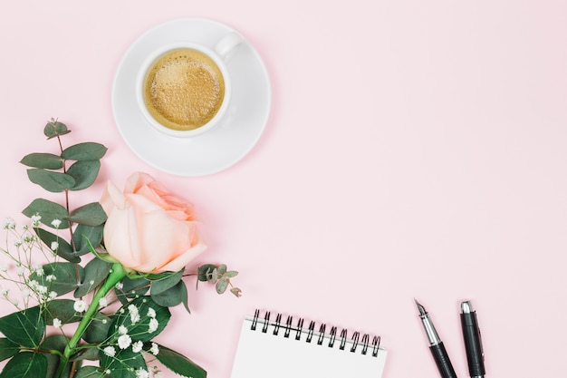 Café; flor rosa; Bloc de notas y pluma sobre fondo rosa