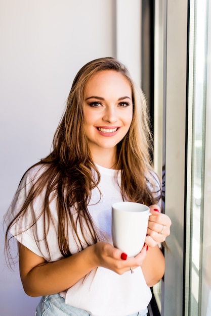 El café de la mañana es mi rutina diaria. Mujer joven beber una taza de café por la mañana cerca de las ventanas en casa mirando a la calle. Humor mañanero.