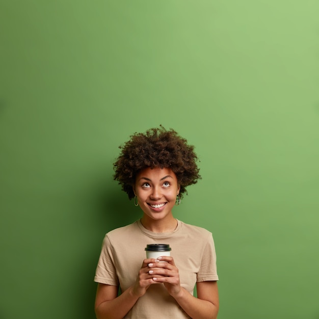 El café nunca es suficiente. Sonriente joven complacida mira hacia arriba en un espacio vacío, sostiene una bebida caliente en un vaso desechable, disfruta de una bebida con cafeína, aislada sobre una pared verde, nota algo arriba