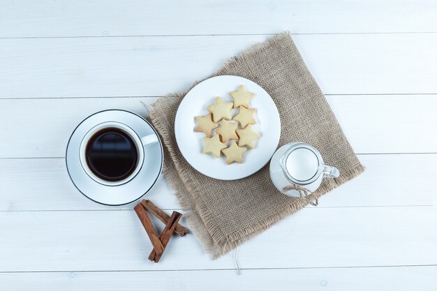 Café en una taza con galletas, canela en rama, vista superior de la leche en madera y un pedazo de fondo de saco