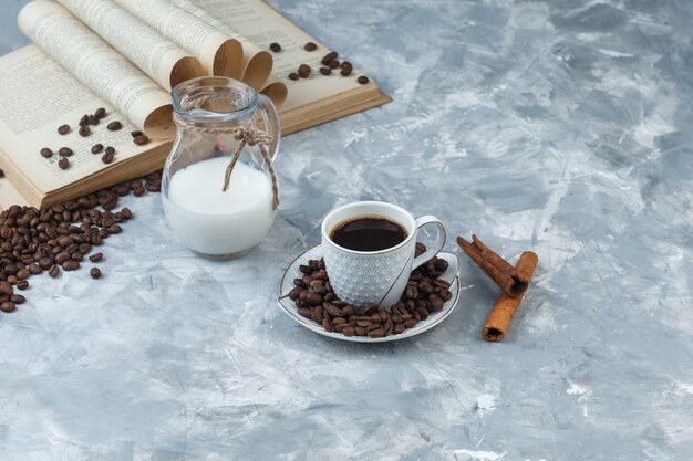 Café en una taza con granos de café, libro, canela en rama, vista de ángulo alto de leche sobre un fondo de yeso gris