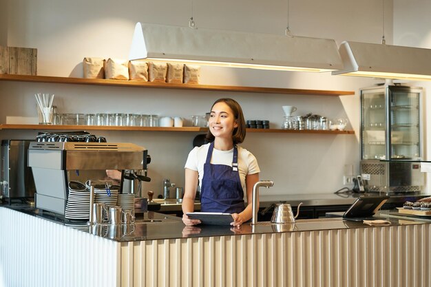 Cafetería sonriente barista coreana trabajando detrás del mostrador usando una tableta para tomar pedidos en el café