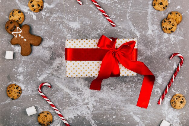 La caja actual con la cinta roja miente en suelo gris con las galletas de la Navidad, los panes de jengibre y los caramelos blancos rojos