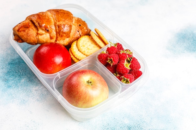 Caja de almuerzo con croissants recién horneados, galletas saladas, frutas y frambuesas.
