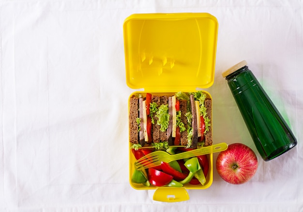 Caja de almuerzo escolar saludable con sandwich de carne y verduras frescas, botella de agua y frutas en la mesa blanca.