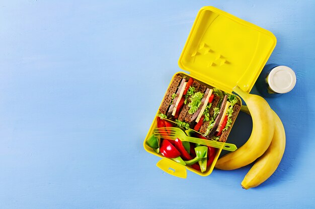 Caja de almuerzo escolar saludable con sandwich de carne y verduras frescas, botella de agua y frutas sobre fondo azul.