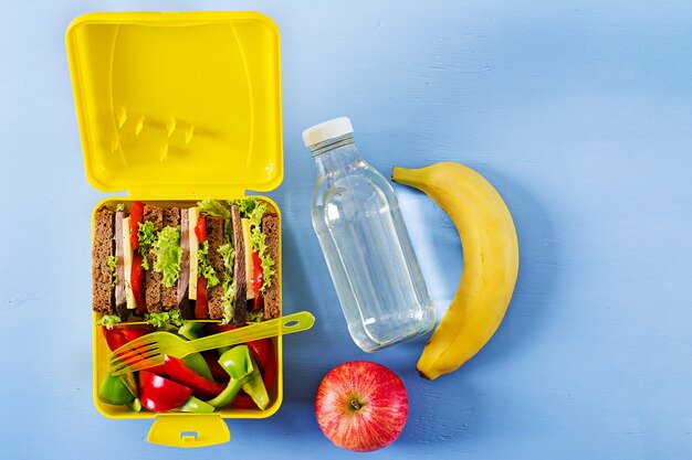 Caja de almuerzo escolar saludable con sándwich de carne y verduras frescas, botella de agua y frutas.