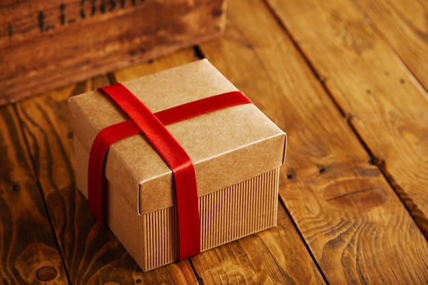 Caja de cartón cuadrada enfocada cerrada y envuelta con cinta de seda roja sobre la mesa de madera rústica junto a la caja vintage