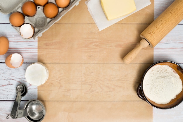 Caja de huevos; mantequilla; rodillo; Harina y cuchara medidora sobre mesa de madera.