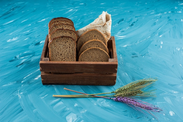 Una caja de madera llena de pan fresco marrón en rodajas con espigas de trigo sobre fondo azul.