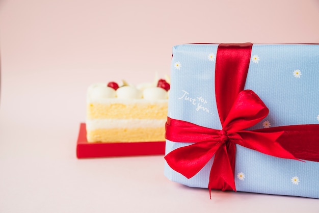 Caja de pastel y regalo envuelto con papel azul y lazo de cinta roja sobre fondo rosa