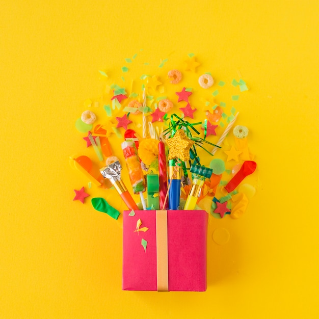 Caja de regalo abierta con dulces y accesorios de fiesta sobre fondo amarillo