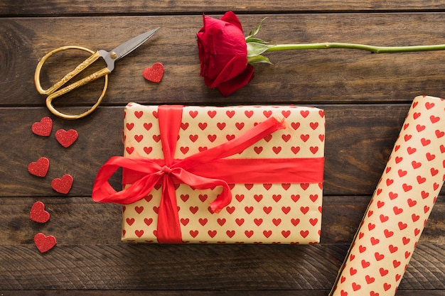 Caja de regalo cerca de rollo de papel cariño, tijeras y flor.