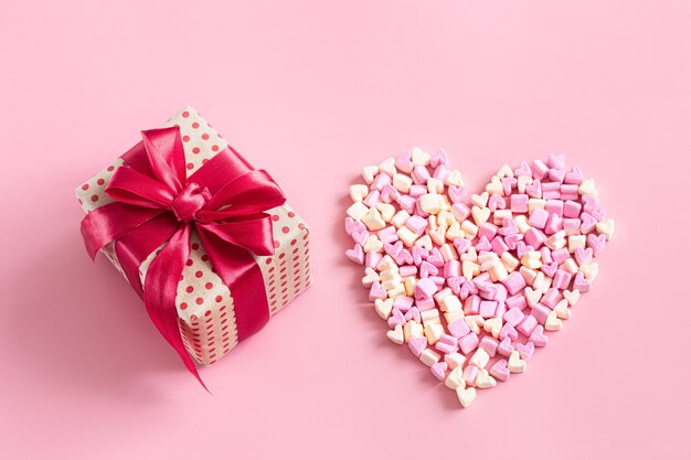 Caja de regalo con lazo rojo y corazón de caramelos en superficie rosa