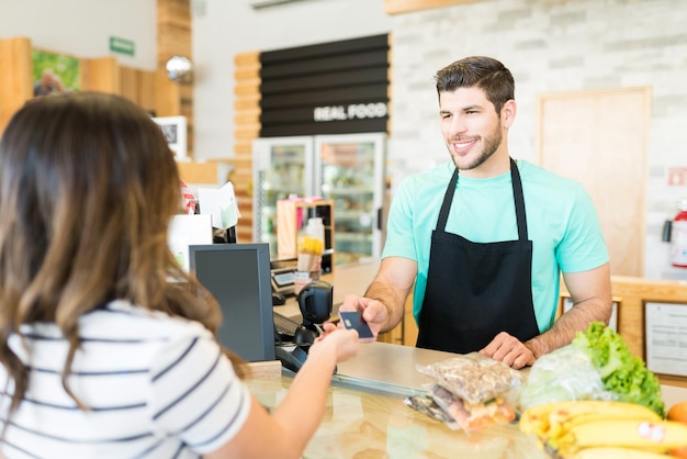 Cajero masculino sonriente que recibe el pago con tarjeta de crédito en el supermercado