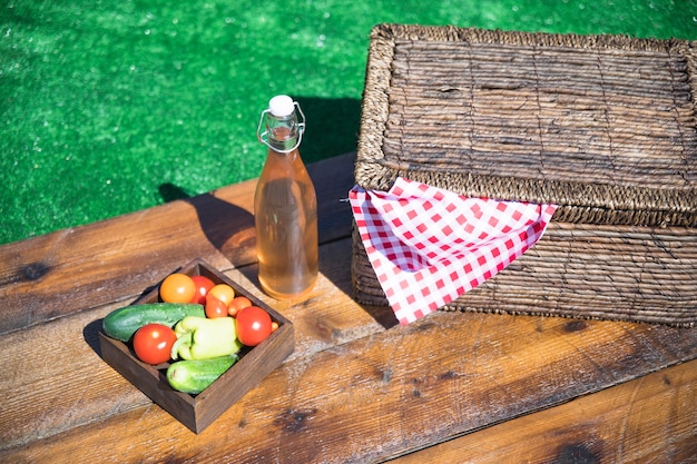 Cajón de verduras; Botella de aceite de oliva y cesta de picnic en mesa de madera.