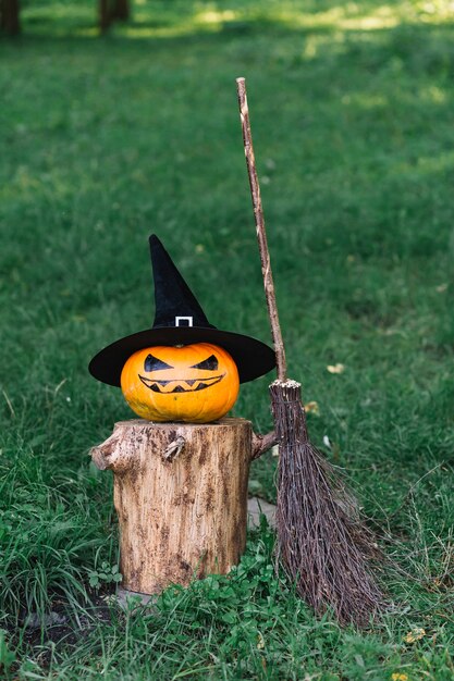 Calabaza de Halloween con sombrero en tocón cerca de escoba