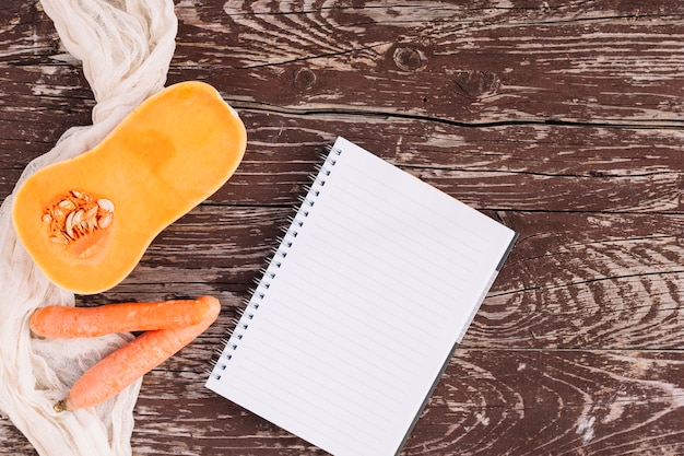 Calabaza a la mitad dividida con dos zanahorias y cuaderno de espiral en una mesa desgastada