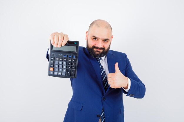 Calculadora de explotación masculina joven, mostrando el pulgar hacia arriba en camisa blanca, chaqueta y mirando confiado. vista frontal.