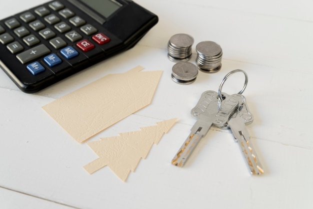 Calculadora; pila de monedas Llaves con casa y árbol recorte de papel en mesa de madera blanca