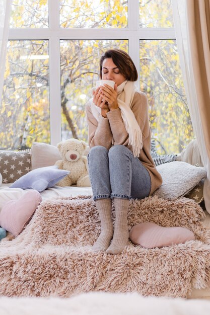 Cálido retrato de mujer sentada en el alféizar de la ventana con una taza de té caliente, café, vistiendo suéter y pañuelo blanco