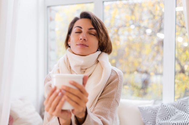 Cálido retrato de mujer sentada en el alféizar de la ventana con una taza de té caliente, café, vistiendo suéter y pañuelo blanco