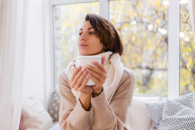 Cálido retrato de mujer sentada en el alféizar de la ventana con una taza de té caliente, café, vistiendo suéter y pañuelo blanco