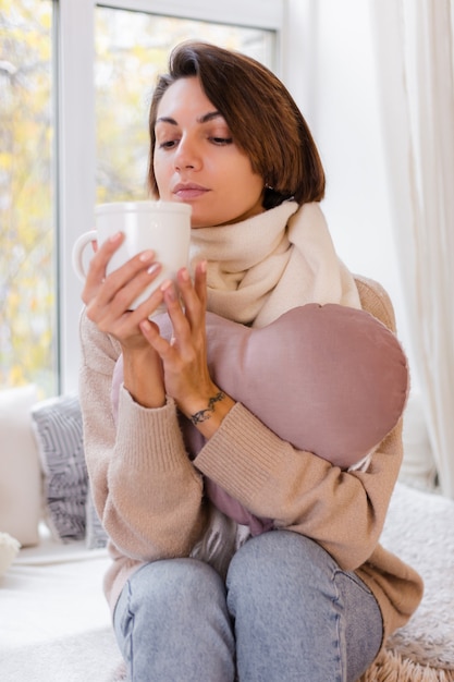 Cálido retrato de mujer sentada en el alféizar de la ventana con una taza de té caliente, café, vistiendo suéter y pañuelo blanco