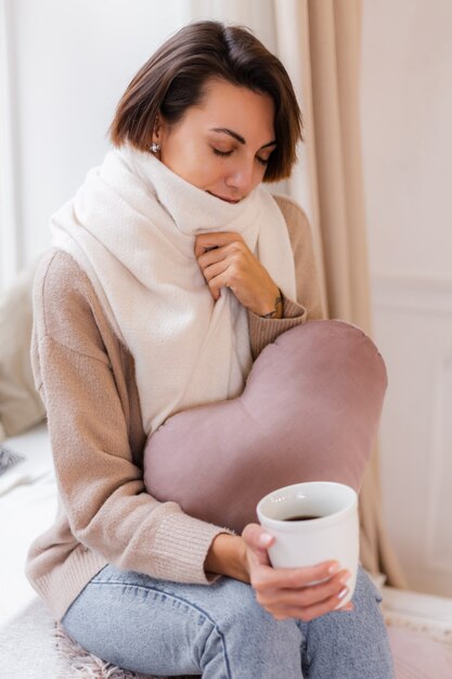 Cálido retrato de mujer sentada en el alféizar de la ventana con una taza de té caliente, café, vistiendo suéter y pañuelo blanco