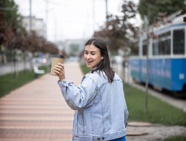 Calle retrato de una mujer joven alegre en un paseo con café en un parque borroso.