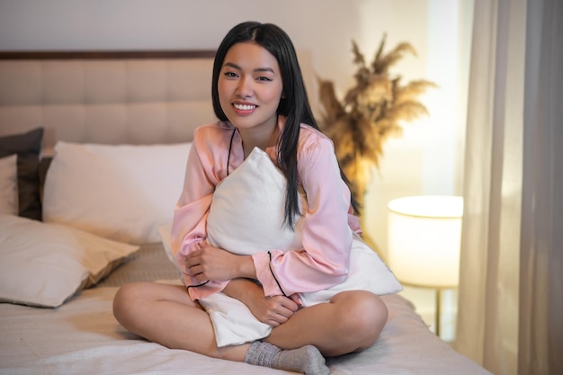 Calma. Adulto joven mujer asiática sonriente con el pelo largo y oscuro en pijama rosa sentado en la cama abrazando la almohada mirando a la cámara