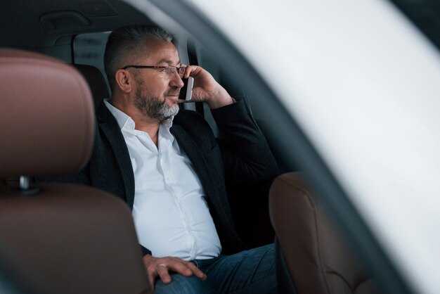 Calma y buen humor. Tener una llamada de negocios mientras está sentado en la parte trasera del automóvil de lujo moderno. Senior hombre con gafas y ropa oficial