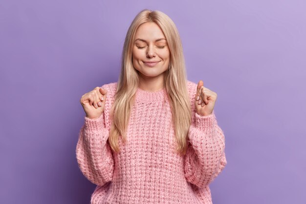 Calma complacida mujer de pelo ligero mantiene los ojos cerrados aprieta los puños espera un momento especial cierra los ojos viste un suéter de punto