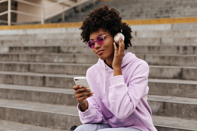 Calma mujer joven relajada en sudadera con capucha púrpura, gafas de sol rosas escucha música en auriculares