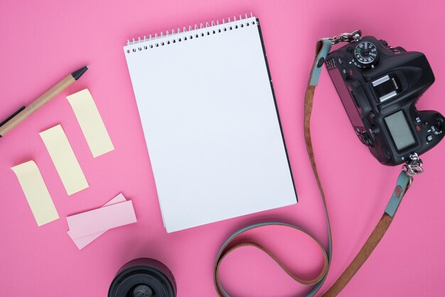 Cámara digital profesional dslr; libreta espiral en blanco; notas adhesivas; bolígrafo; lente de cámara y cinturón sobre fondo rosa