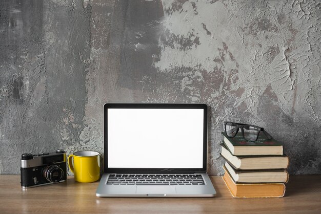 Cámara; vaso; libros apilados; Gafas y laptop con pantalla blanca en blanco en tablero de madera.