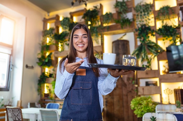 Camarera feliz sosteniendo una bandeja con una taza de café trabajando en la cafetería y sirviendo la mesa