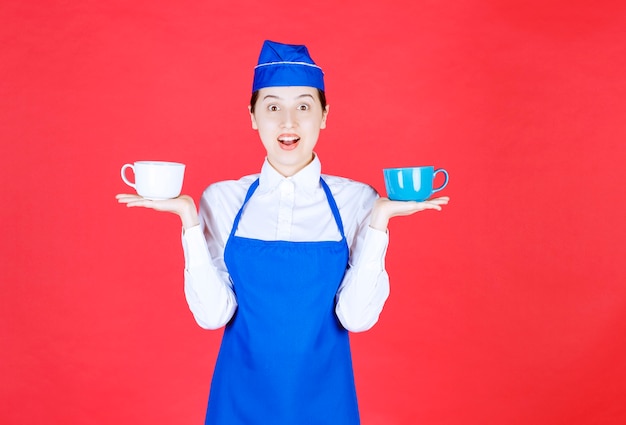 Camarera de mujer en uniforme de pie y sosteniendo tazas de colores en la pared roja.