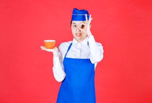 Camarera de mujer en uniforme sosteniendo un cuenco naranja y haciendo un gesto bien cerca del ojo.