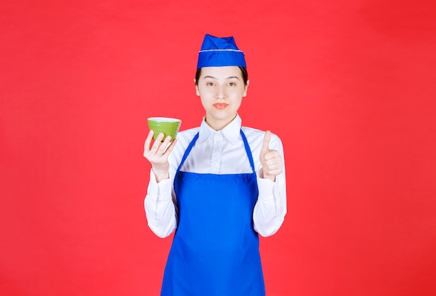 Camarera de mujer en uniforme sosteniendo un cuenco verde y mostrando el pulgar hacia arriba.