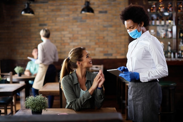 Camarera negra feliz con mascarilla protectora mientras toma el pedido del cliente en un panel táctil