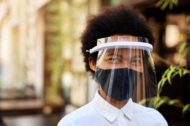 Camarera negra feliz con visera y mascarilla protectora en un café al aire libre