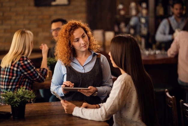 Camarera pelirroja hablando con una mujer y escribiendo el pedido en una tableta digital en un pub