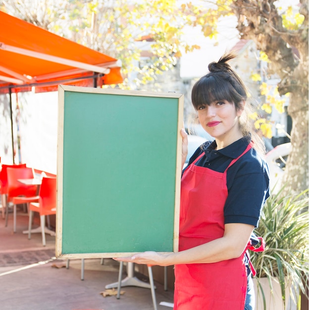 Camarera de sexo femenino que sostiene el tablero verde en blanco del menú en el café al aire libre