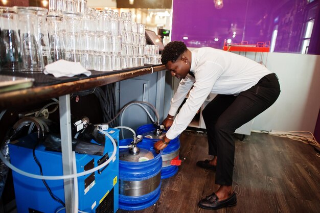 Camarero afroamericano en el bar con barril de cerveza de acero inoxidable
